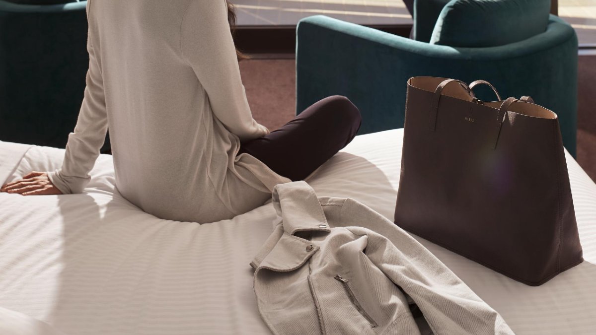 A woman sits on the edge of a bed with a brown leather handbag next to her