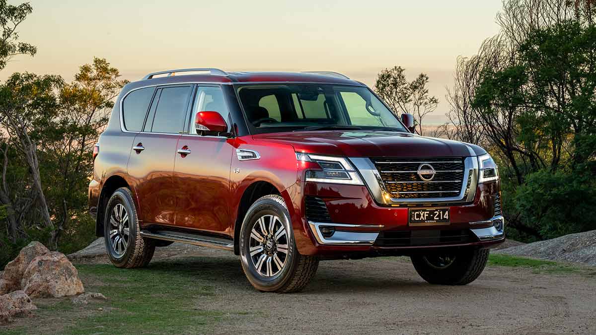 Red Nissan Patrol SUV parked in coastal camping spot lined with granite rocks and shrub.