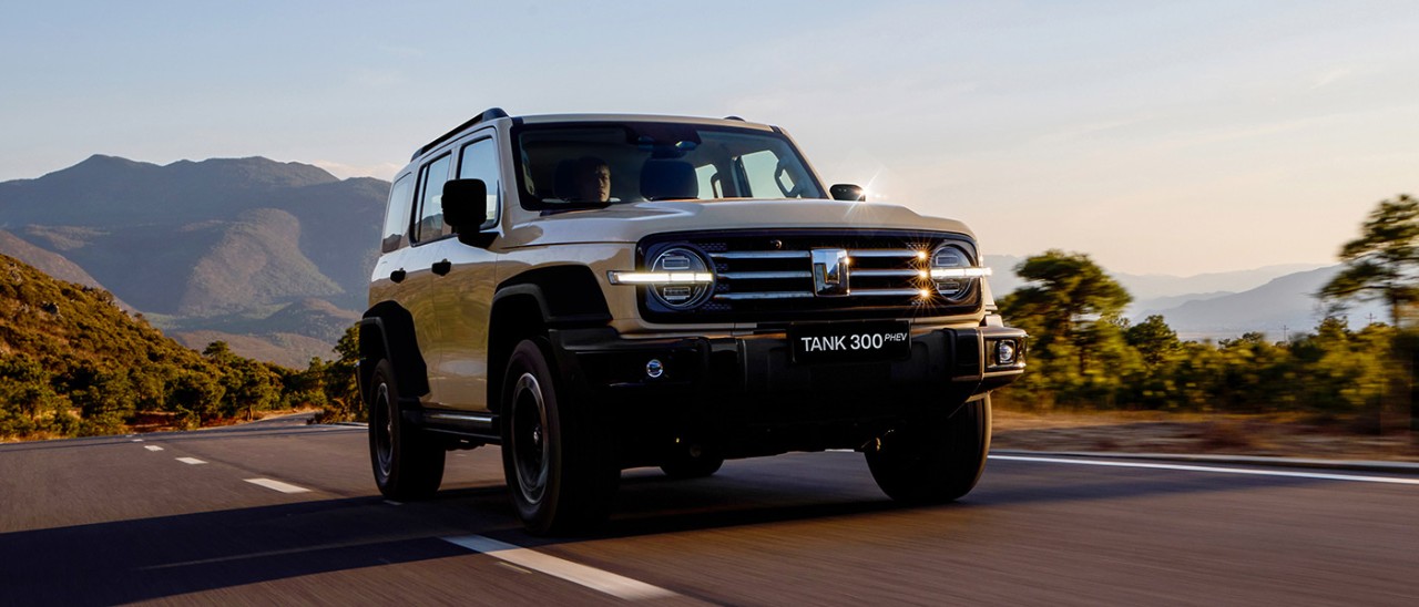 Cream-coloured GWM Tank 300 SUV driving on bitumen road in mountainous area with sunspots on 3-bar chrome grille.
