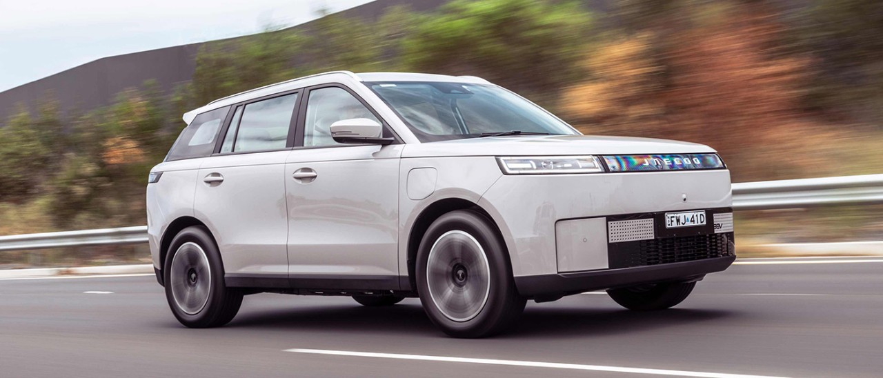 White Jaecoo J5 EV small SUV on freeway in left lane next to armco, small trees and tall grey sound barrier.