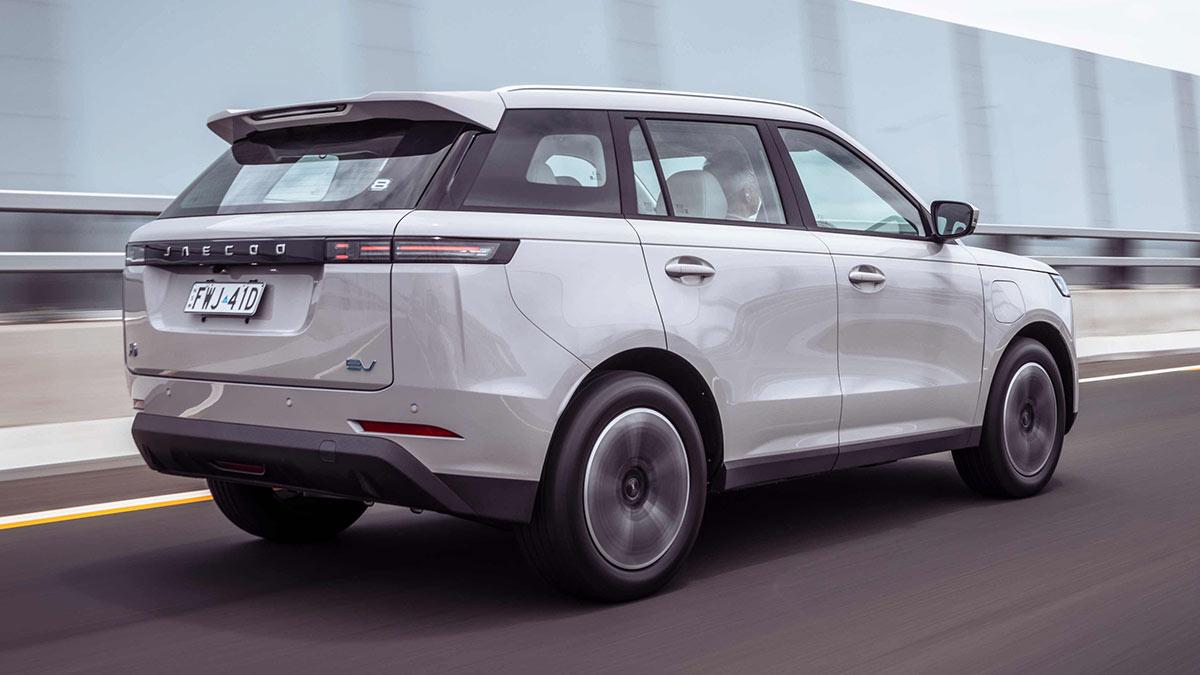 Rear view of white Jaecoo J5 EV on freeway with concrete barrier, guard rails and transparent noise barrier panels at side..
