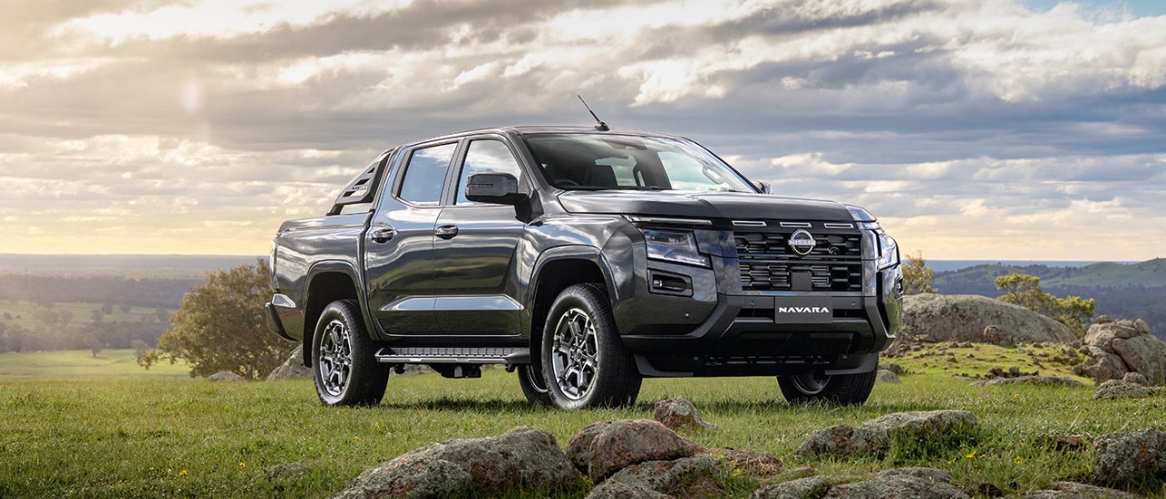 Black Nissan Navara ST dual cab ute on grassy, rocky hilltop in rural area with bright blue sky and clouds.