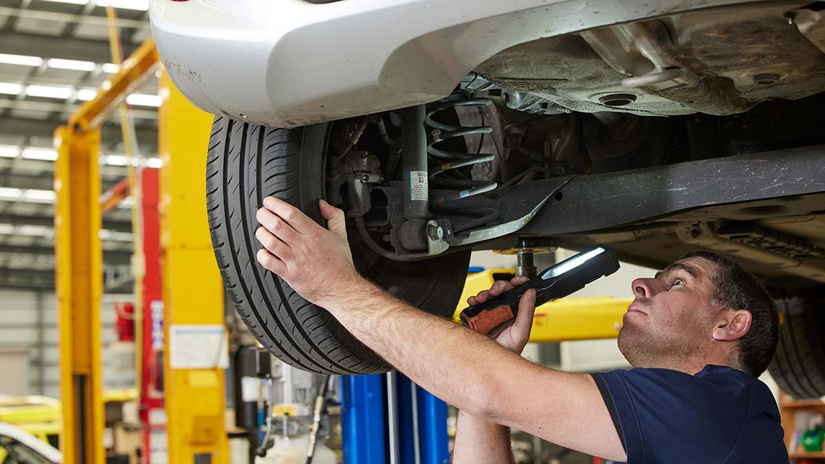 Car mechanic under hoist in workshop inspecting car's suspension with torch