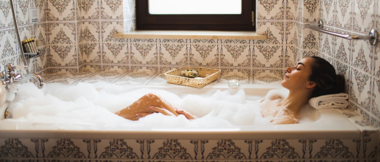 A woman enjoying a bubble bath in an ornately tiled bathroom. She is resting her head on a folded pillow and tea lights are on the bath ledge