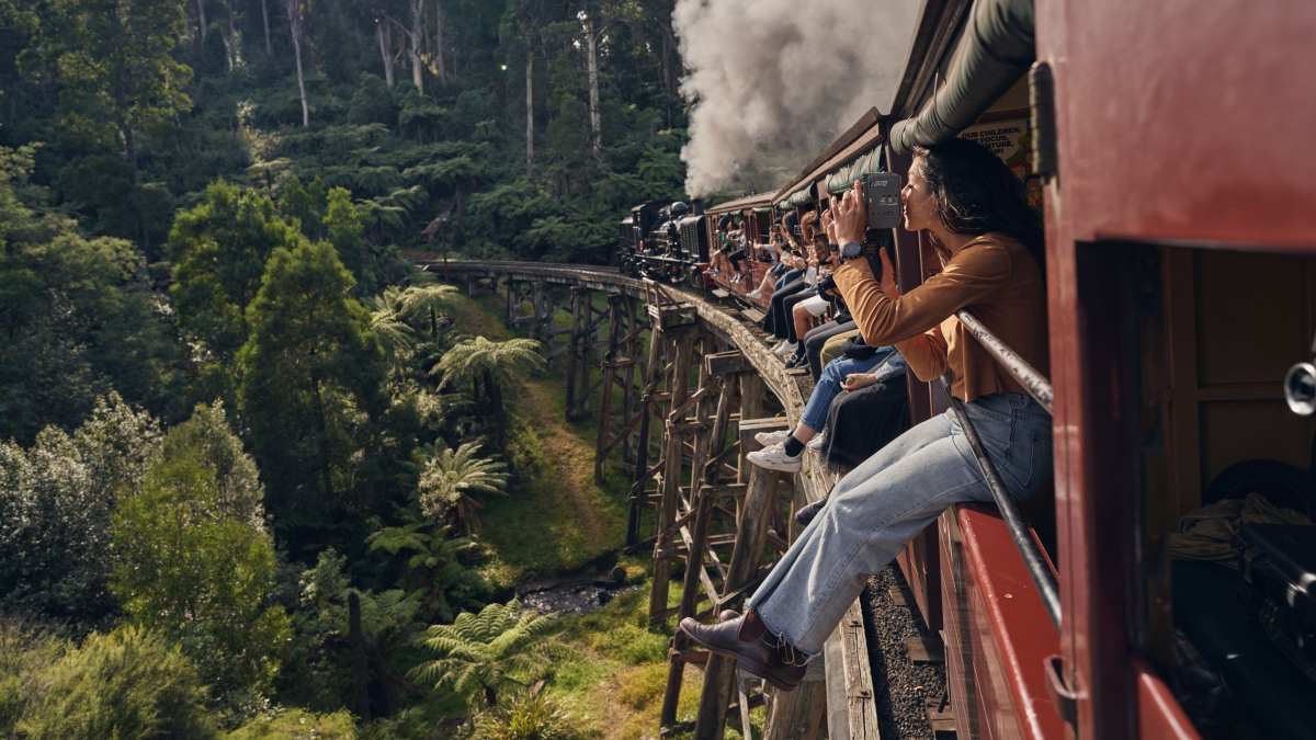People sitting on the sills as steam train Puffing Billy travels over a wooden bridge