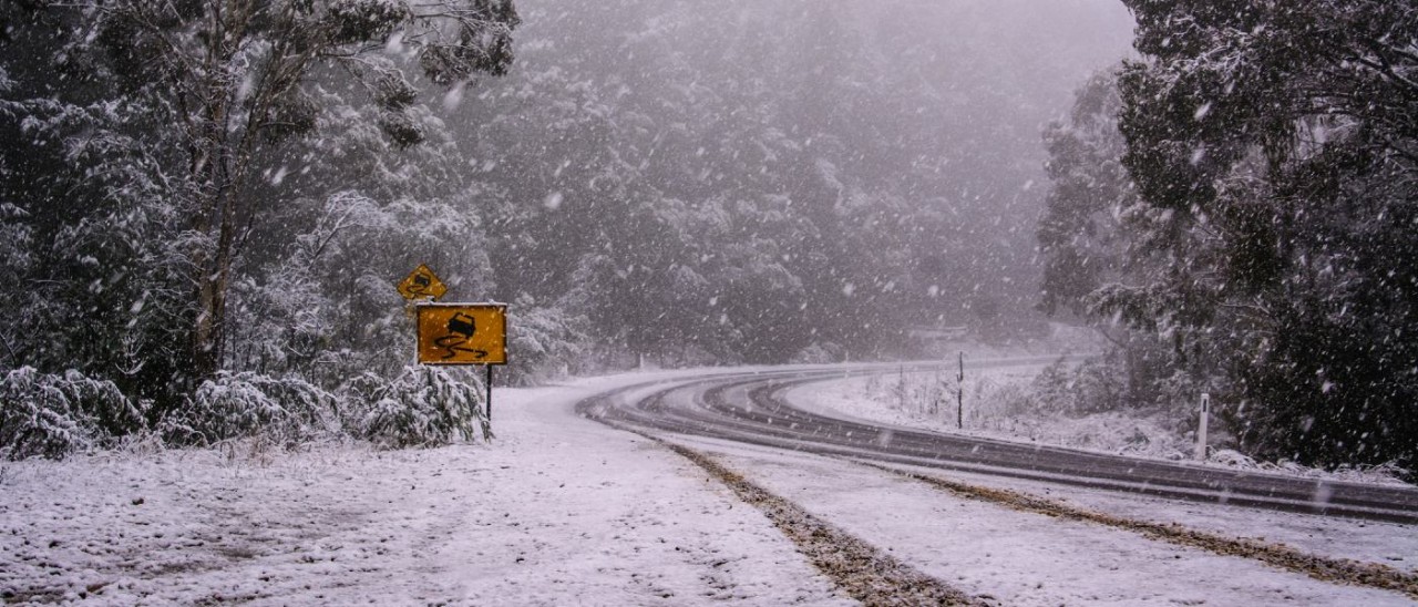 A snowy mountain road in Australia. A yellow road sign warning of slippery roads can be seen on the left.