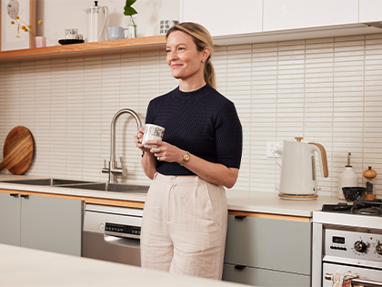 Woman holding a mug while standing in her kitchen amongst appliances.