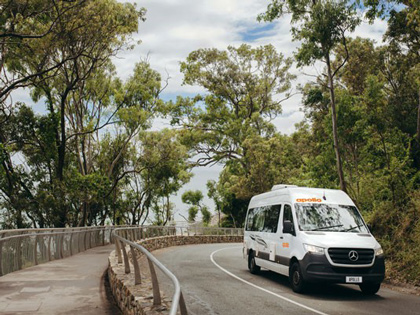 A white campervan driving with trees and an ocean background.