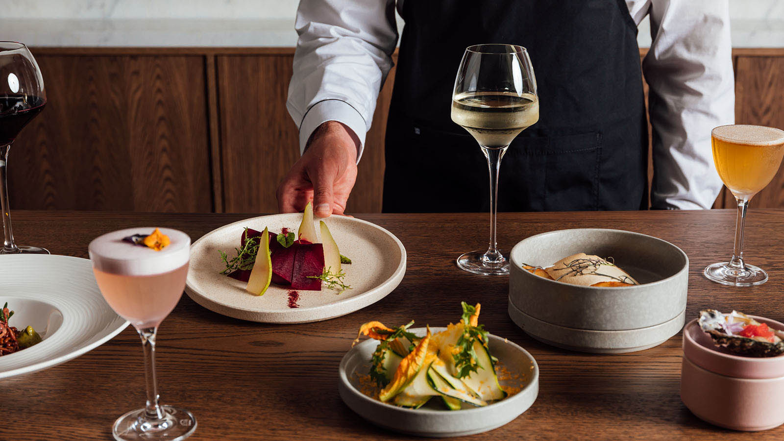 A selection of small dishes, a glass of white wine and two cocktails displayed on a wooden table at RACV City Club's Wine Bar.