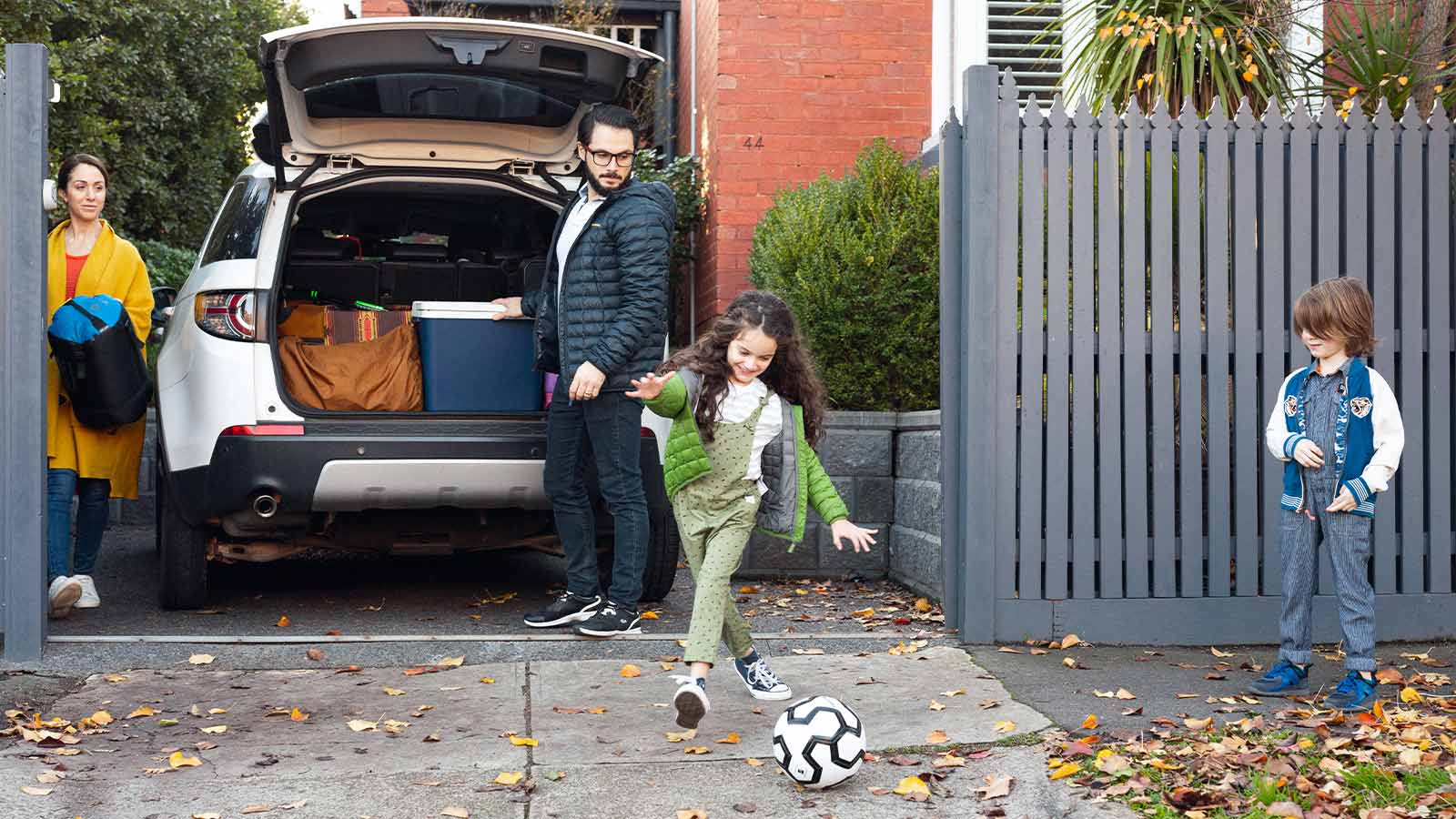 Parents watching their child kick a soccer ball in their driveway, while getting ready to go on a holiday.