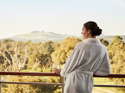 Person wearing a bathrobe and holding a glass while looking out from a balcony at RACV Goldfields Resort.