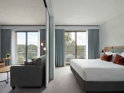 Goldfields studio room with a large bed, a sofa, and a table. The room has two large windows with curtains and a view of greenery outside.