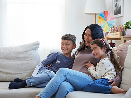 Parent and two kids sitting and smiling together on a grey couch in a living room.
