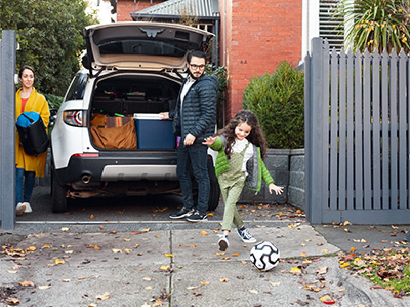 Parents watching their child kick a soccer ball in their driveway, while getting ready to go on a holiday. 