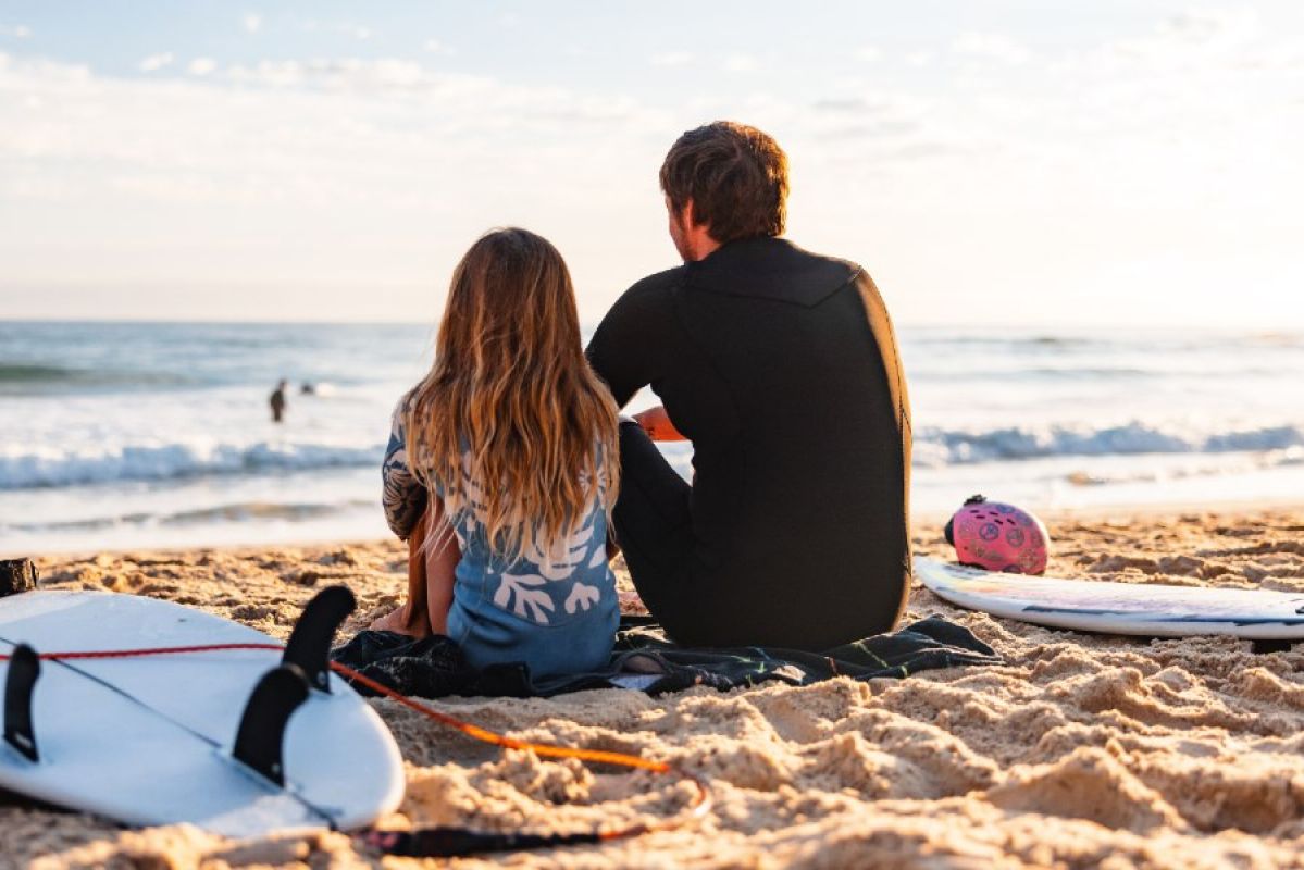 father and daughter sitting on beach with surfboards looking out to sea