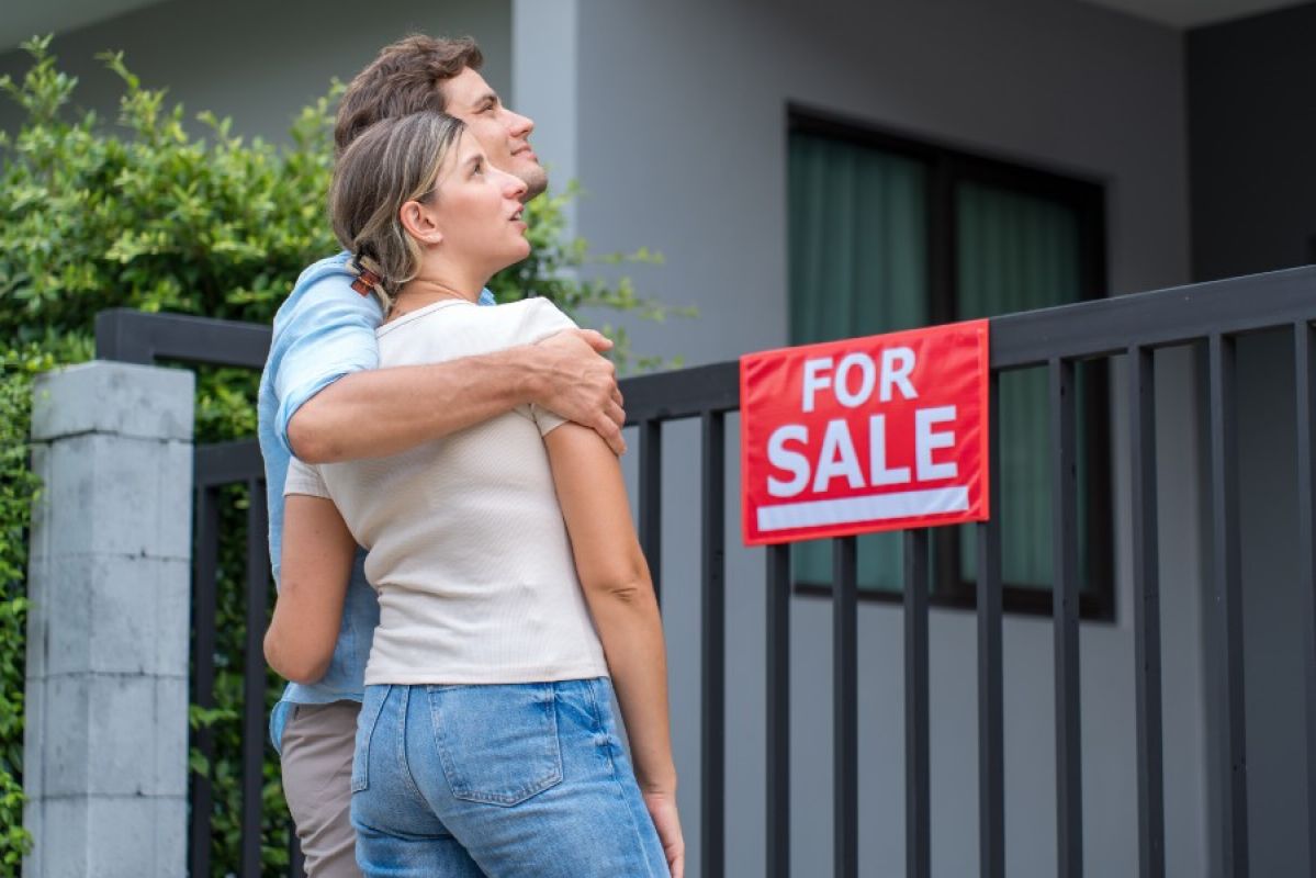 young couple hugging and looking at a home with a for sale sign
