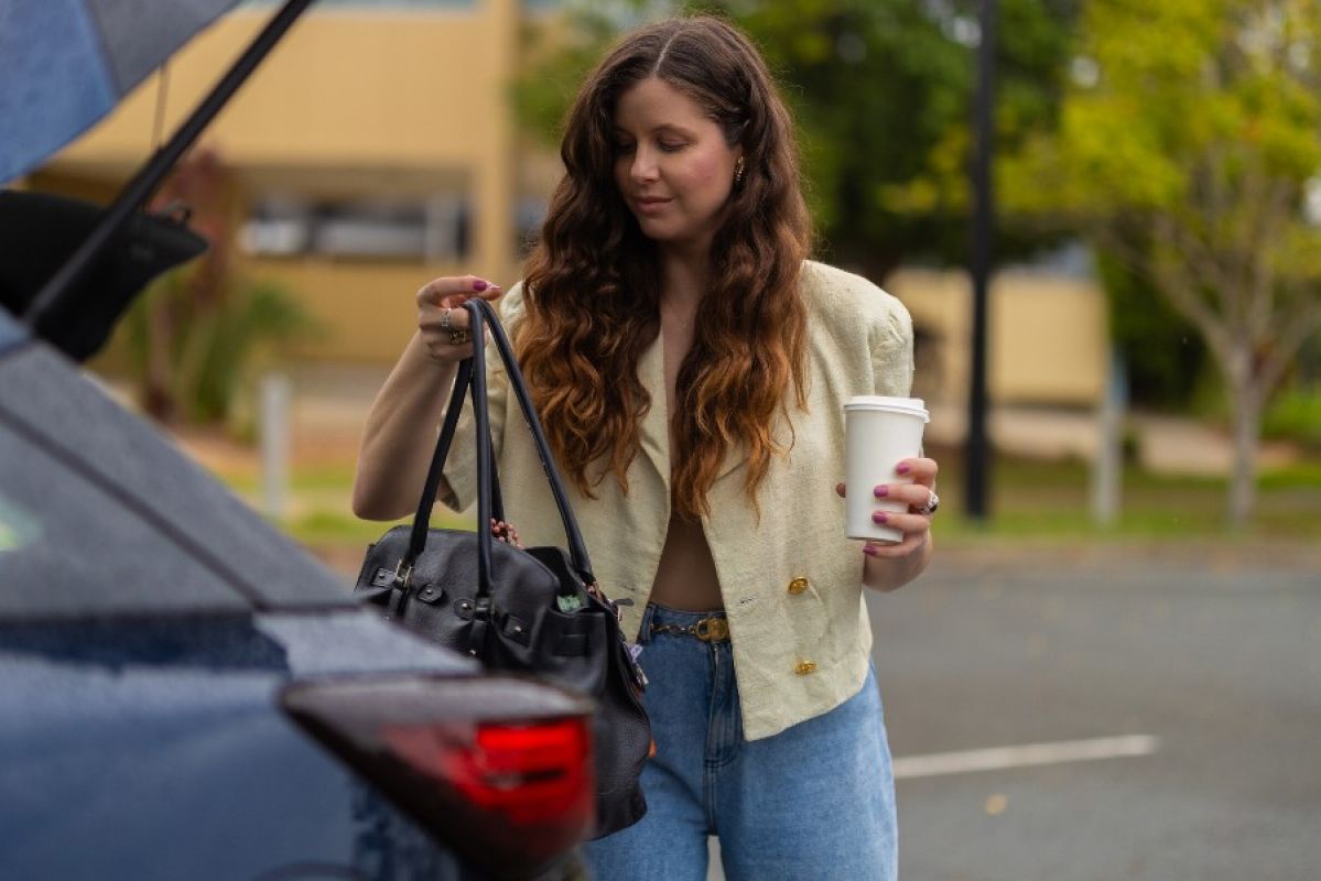 A young woman taking a stylish black leather handbag out of the boot of her car. She is holding a takeaway coffee in her other hand