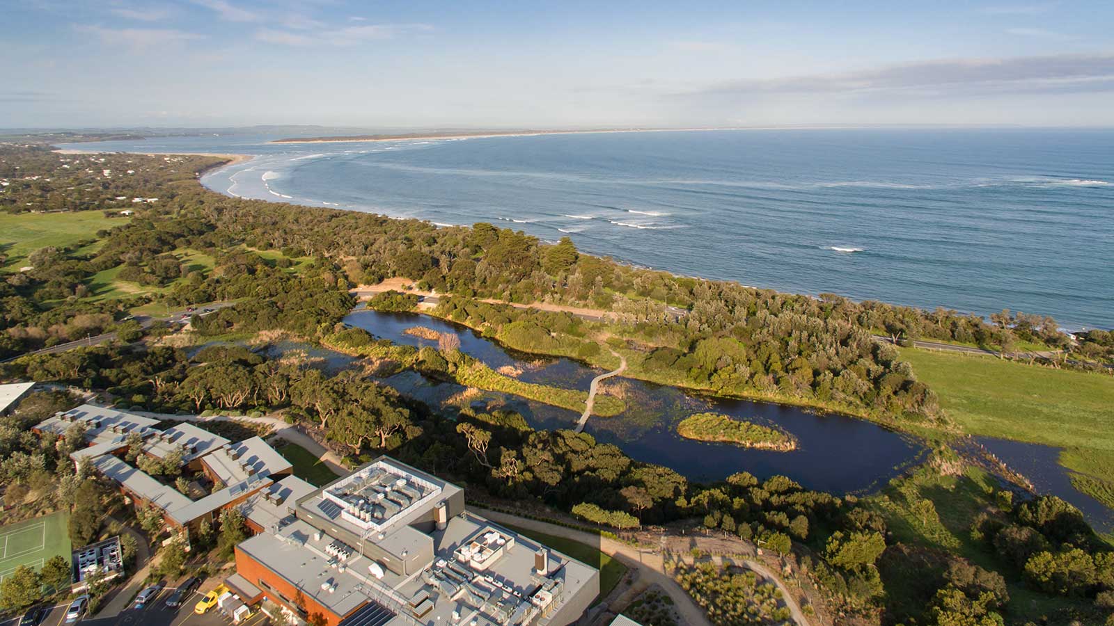RACV Inverloch Resort on a sunny day, in front of green trees and fields, and the beach.