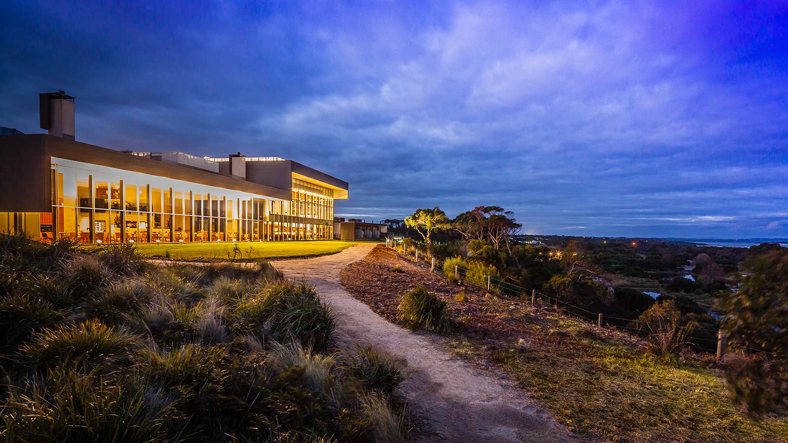 Path surrounded by greenery leading to the main RACV Inverloch Resort building, lit up at night.