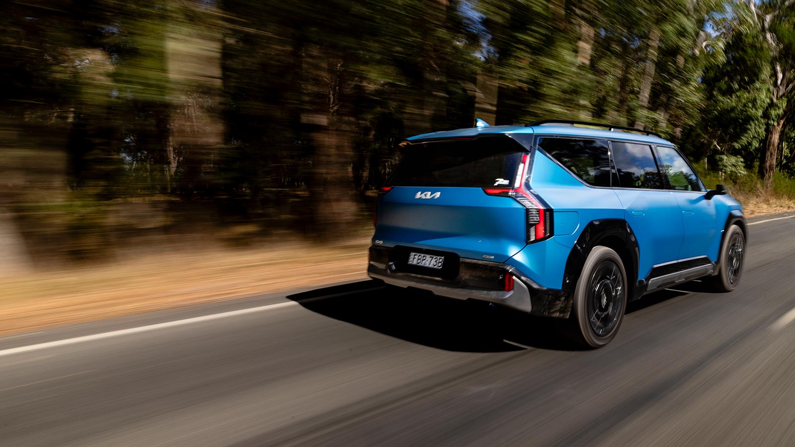 A blue Kia EV9 SUV driving on the left side of a road