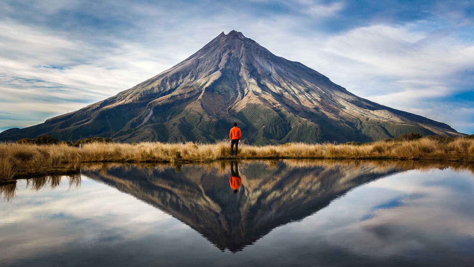 Man standing infront of Mount Taranaki in New Zealand with the reflection showing in the lake.