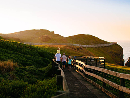 Parents and two kids walking down wooden bridge surrounded by greenery at sunset.