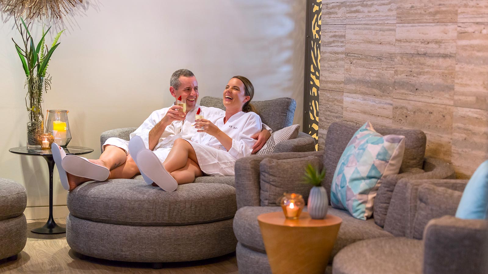 Couple laughing together in bathrobes in the relaxation lounge at RACV Noosa Resort's One Spa, with drinks in their hands.