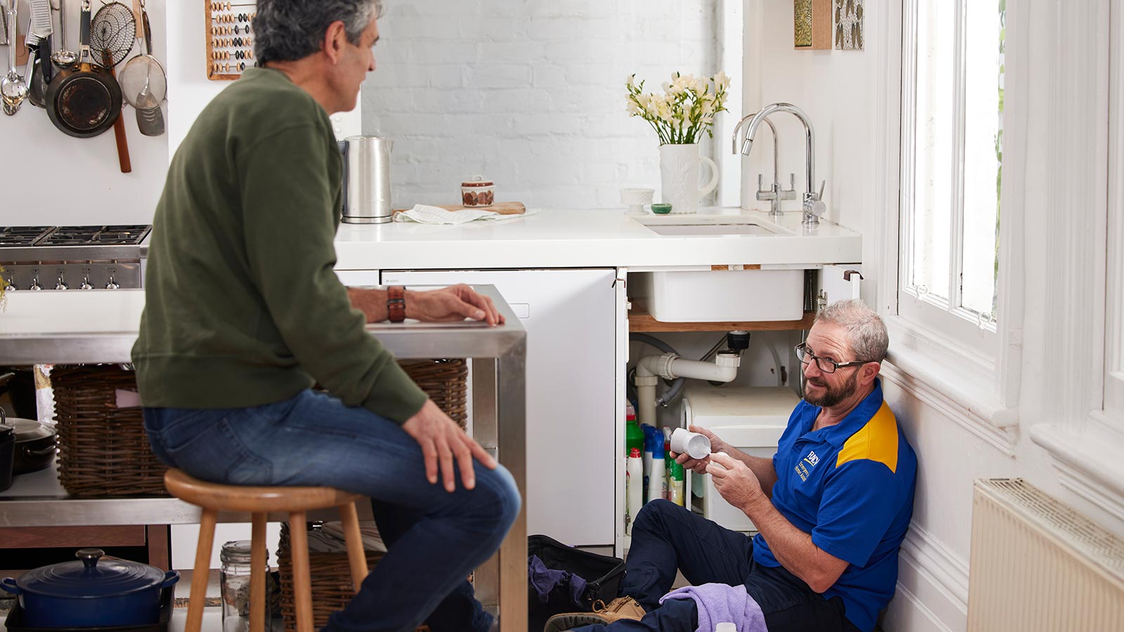 Plumber sitting on the ground fixing kitchen sink while talking to the homeowner.