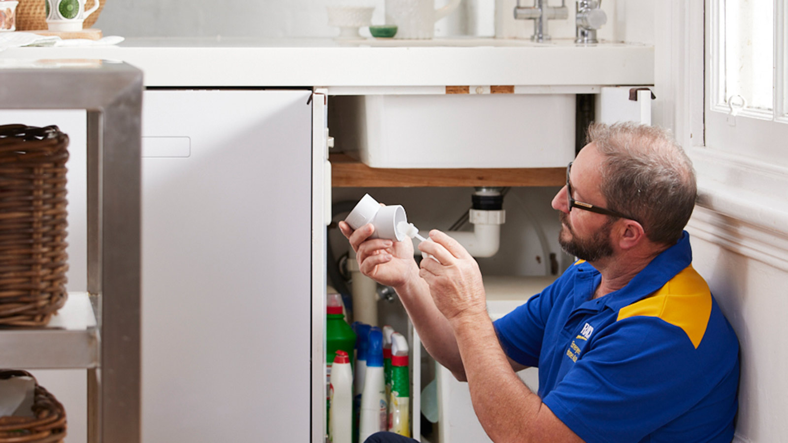 Plumber working on pipes whilst sitting next to kitchen sink.