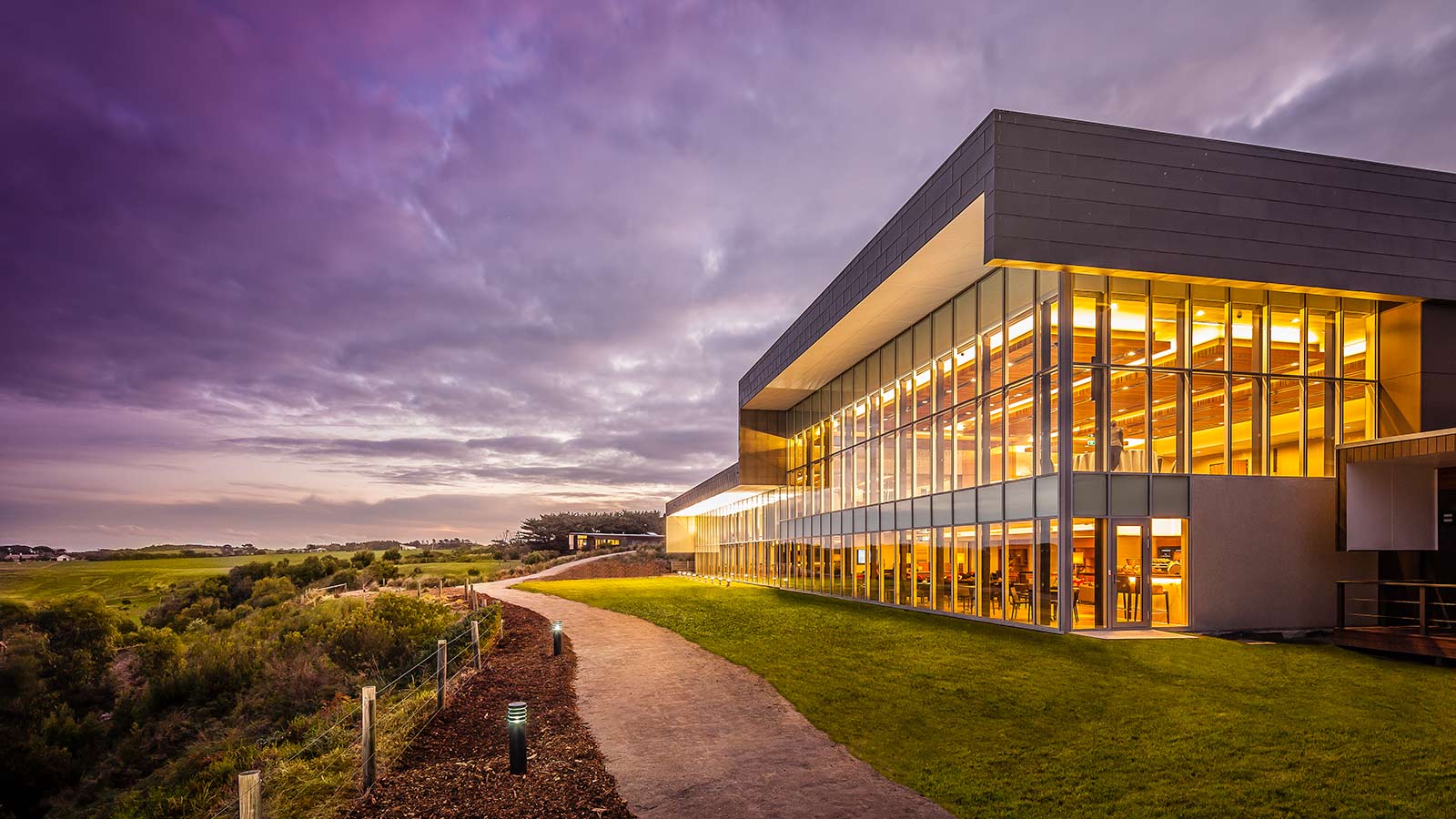 Path surrounded by greenery leading to the main RACV Inverloch Resort building, lit up at dusk.