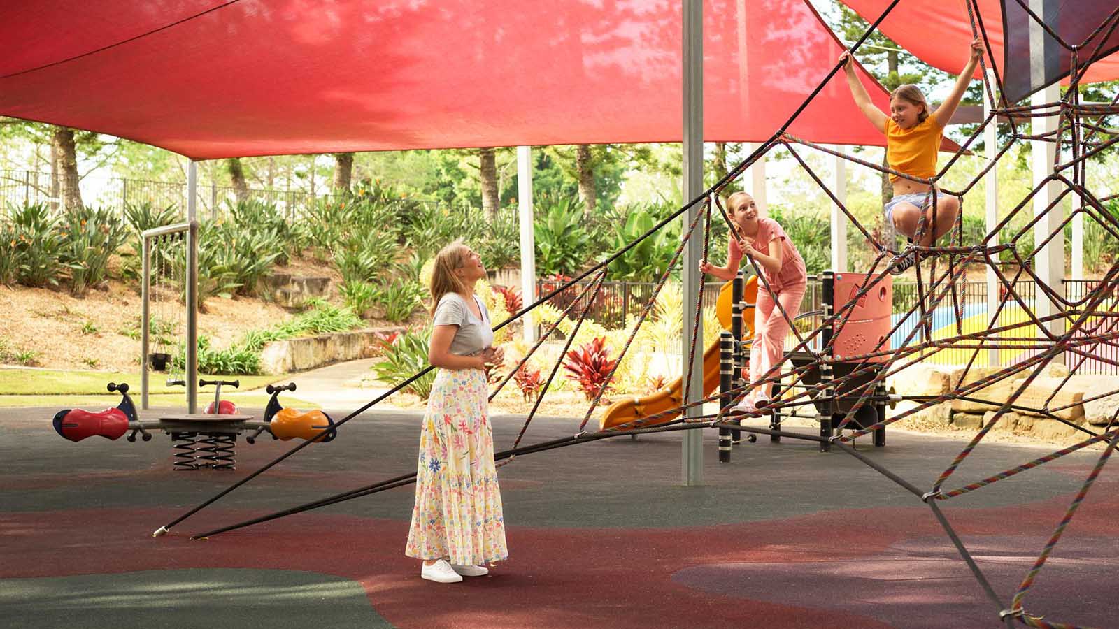 Mother smiling and overlooking her two children playing on a spider web climber on the Royal Pines kids playground.