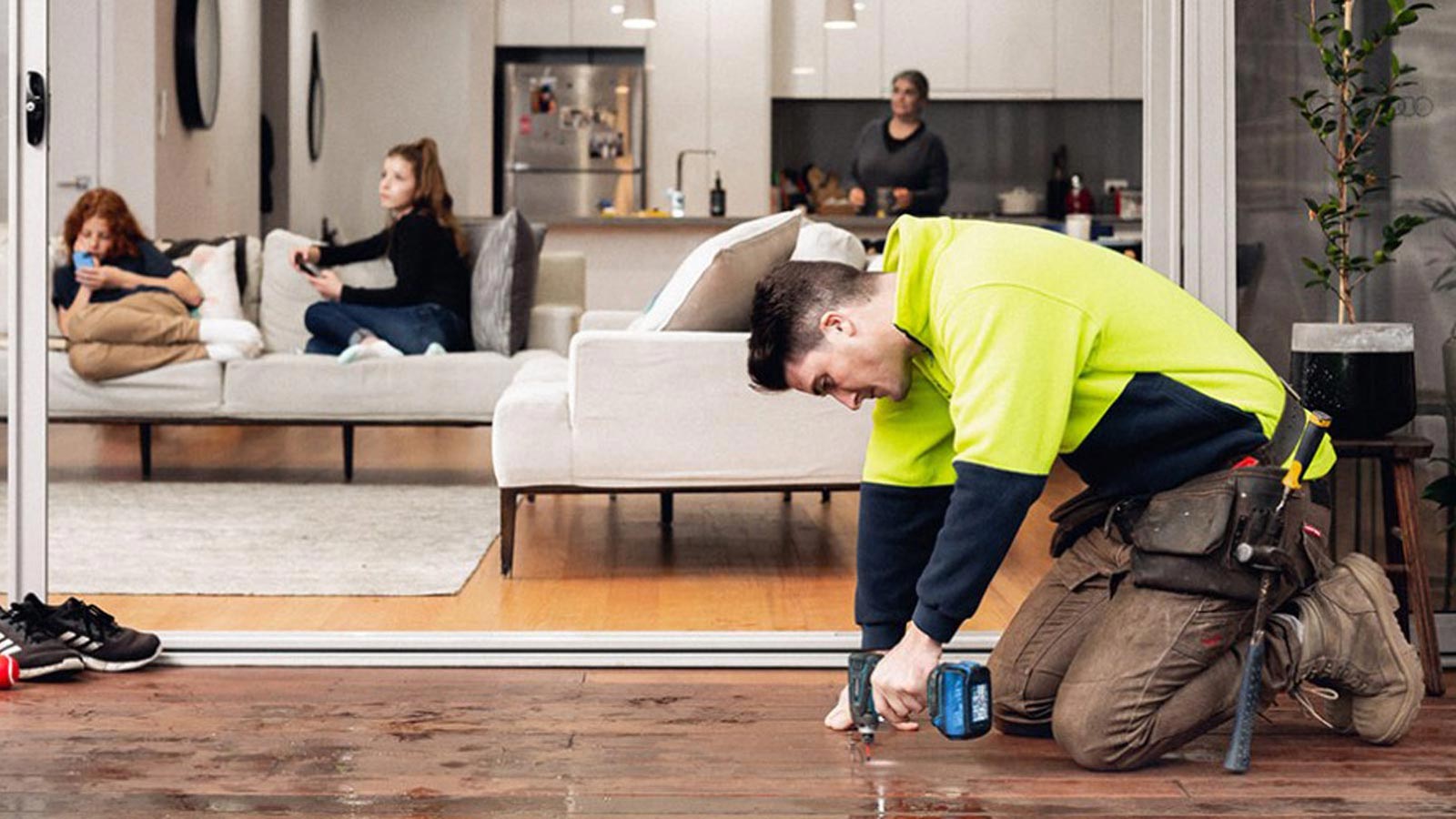Tradesman repairing decking whilst two teenagers and mother are watching television from the inside open plan living area.