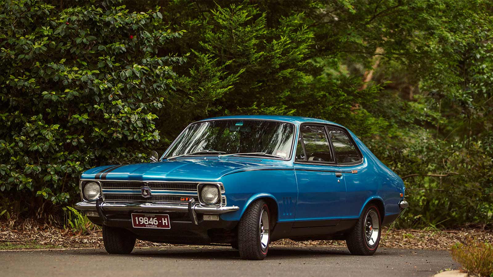 Blue antique Holden Monaro parked outside in front of greenery.