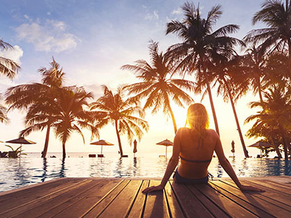 Silhouette of woman lounging in front of pool and palm trees at sunset.